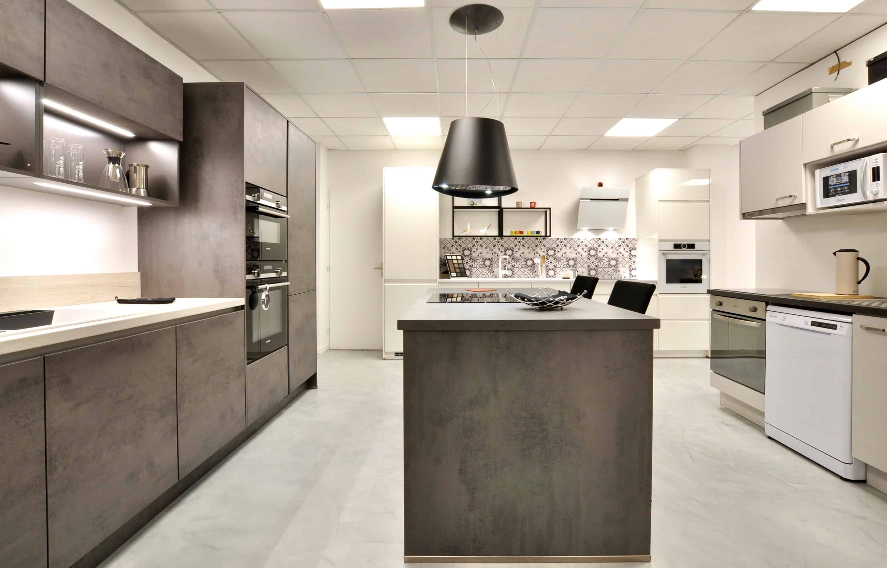 Exposition îlot + linéaire promotion immobilière Façade et plan de travail en décor béton gris ardoise, électroménager Siemens, hotte suspendue Elica. A droite, linéaire promotion immobilière avec façades en finition gris soie, électroménager Candy, évier et robinetterie Blanco.